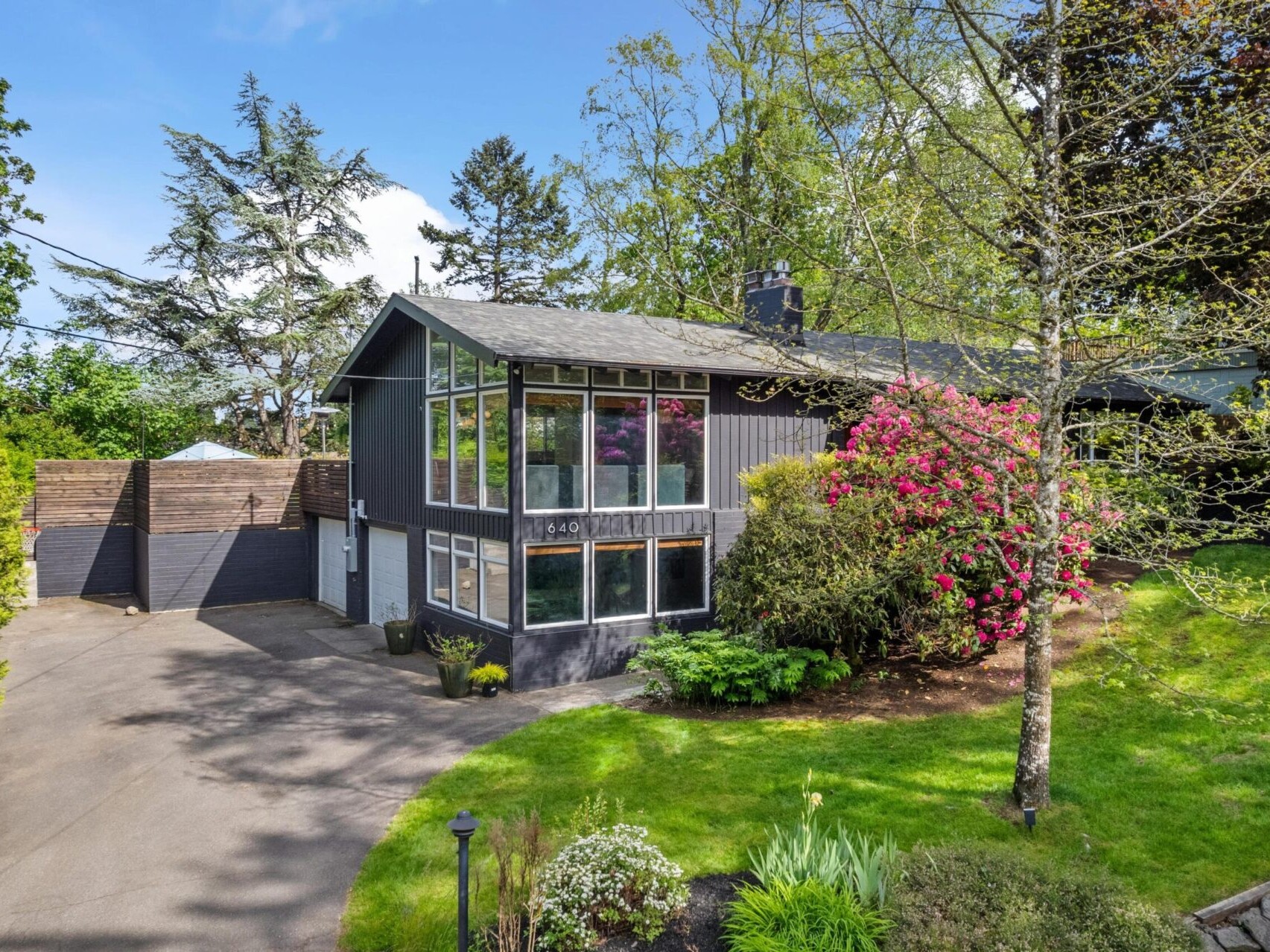 A modern, two-story house with large windows, a gray exterior, and a sloped roof. Its surrounded by lush greenery and colorful flowering bushes with a paved driveway on a sunny day.