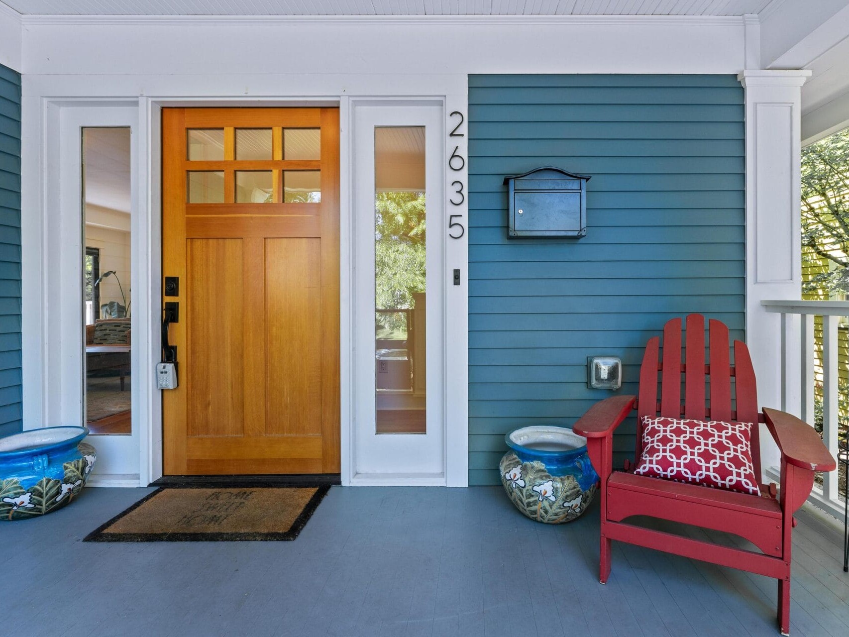 Front porch of a house with a wooden door, house number 2635 displayed vertically. A red Adirondack chair with a patterned cushion sits on the right, flanked by two decorative potted plants. A black mailbox is mounted on the wall.