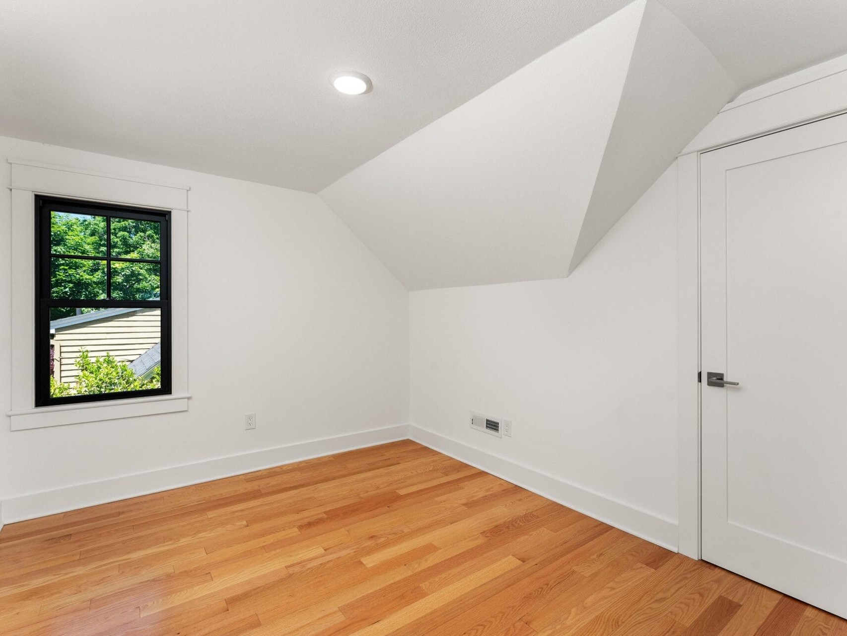 Small empty room with sloped ceiling, white walls, and wooden floor. A single window with black trim shows greenery outside. A closed white door is on the right. A ceiling light illuminates the space.