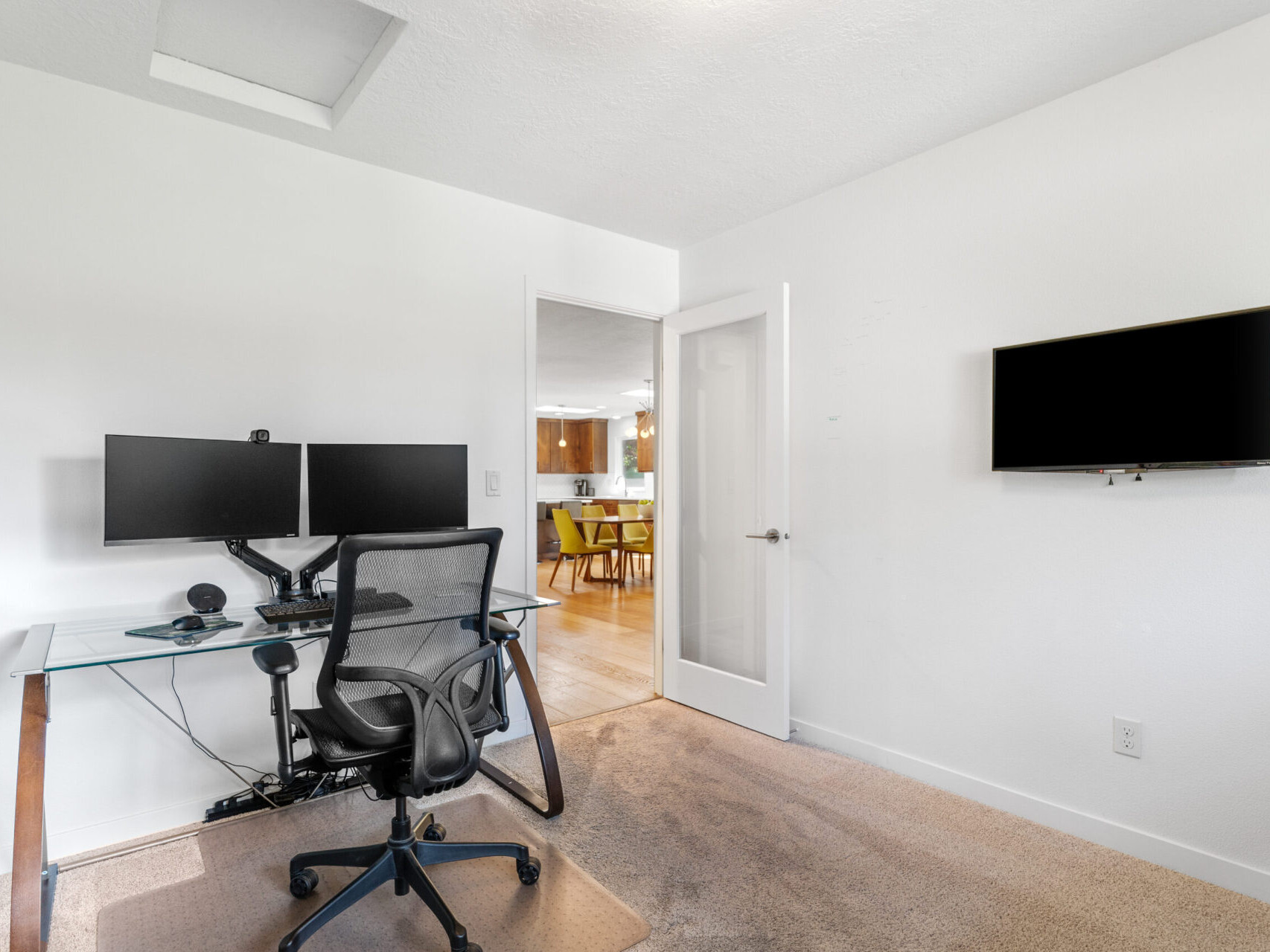 A home office with a glass desk, dual monitors, and a swivel chair. A wall-mounted TV is on the right, and a doorway opens to a living area with a dining table visible in the background. The room has white walls and beige carpet.