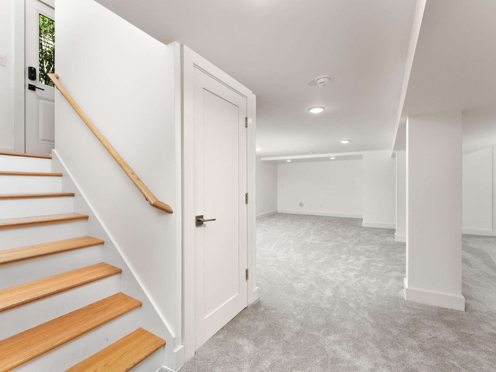 A newly renovated, carpeted basement with white walls. The space features a staircase with wooden steps leading upstairs. There are closed white doors and recessed lighting in the ceiling, creating a bright and clean atmosphere.