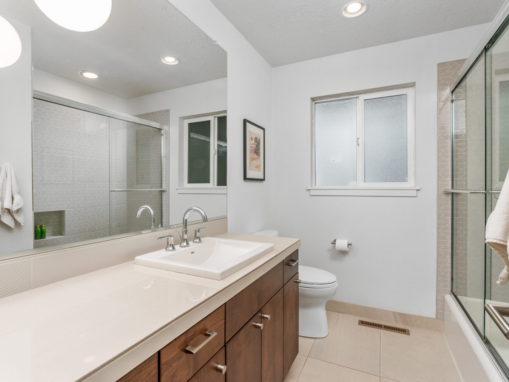 A modern bathroom with a large mirror above a wide sink counter and wooden cabinets. There is a glass-enclosed shower, a toilet, and a frosted window. Recessed lighting brightens the space. A towel hangs on the wall.