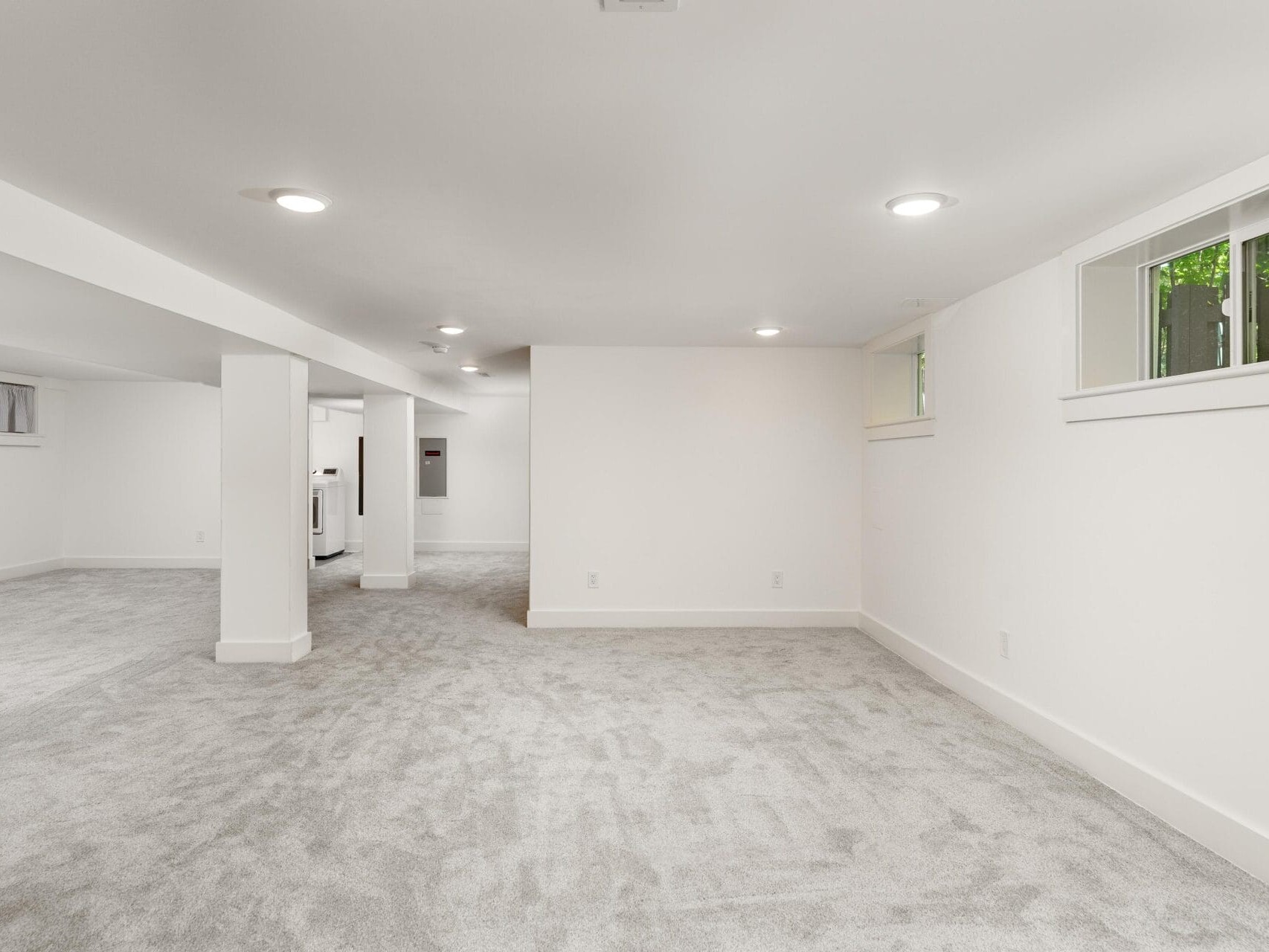 A spacious, unfinished basement with carpeted floors and white walls. The ceiling features recessed lighting, and there are small rectangular windows along the top of the walls. The area appears open and versatile.