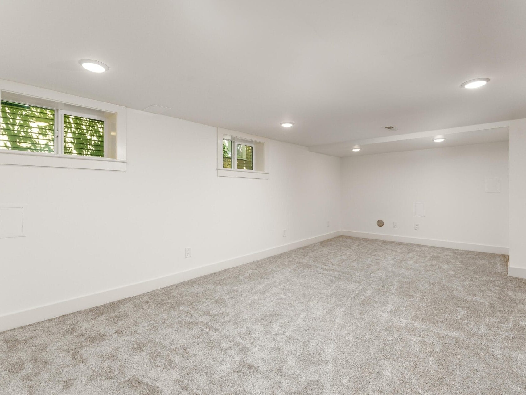 A spacious, empty basement with white walls and light gray carpet. There are small, rectangular windows near the ceiling on one wall, letting in natural light. Recessed lighting fixtures illuminate the room.