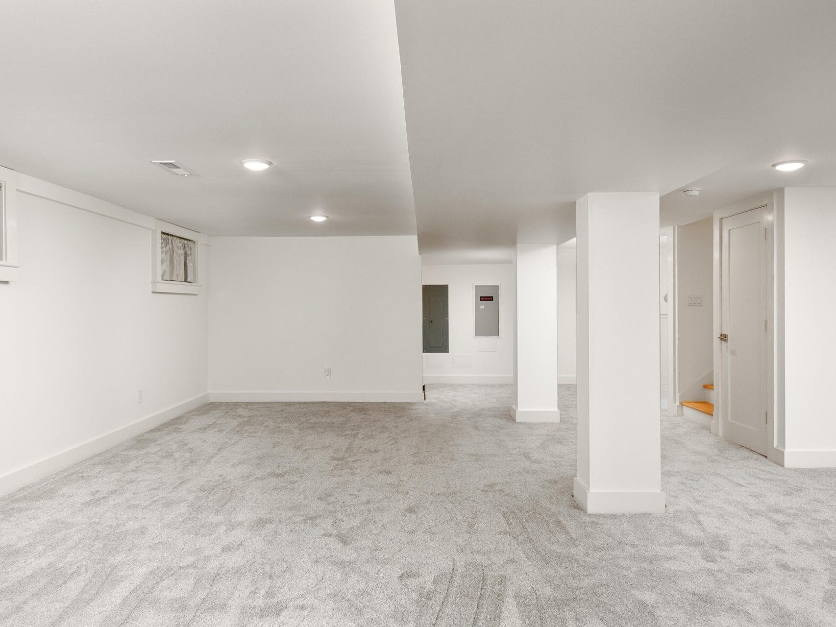 A spacious, empty basement with light gray carpet and white walls. The room has small windows near the ceiling, recessed lighting, and a visible electrical panel. A staircase with an orange-colored step is partially visible on the right.