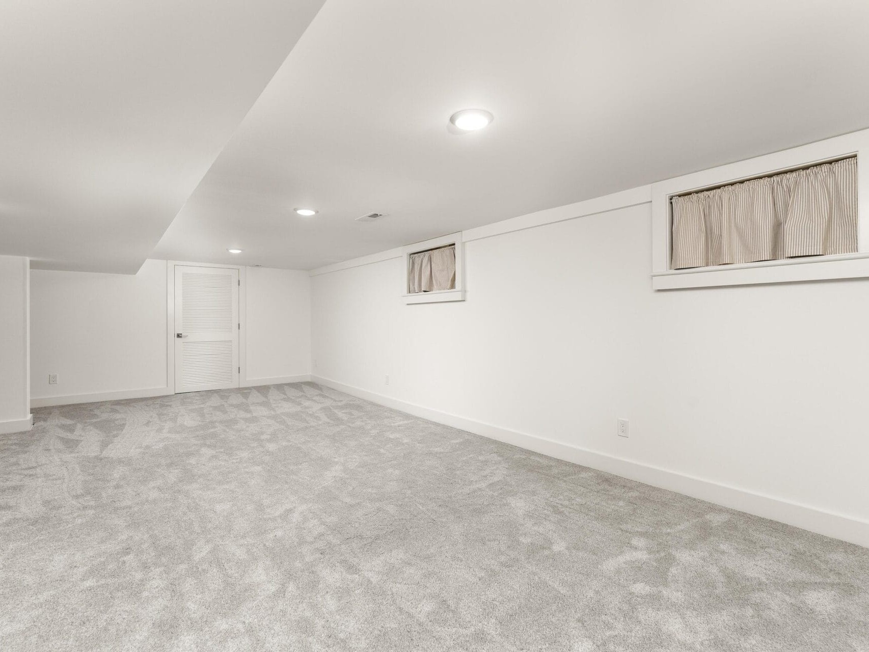 A spacious, empty basement room with light gray carpet and white walls. Two small windows with curtains are set high on the wall. Recessed lights brighten the room, and a closed white door is at the far end.