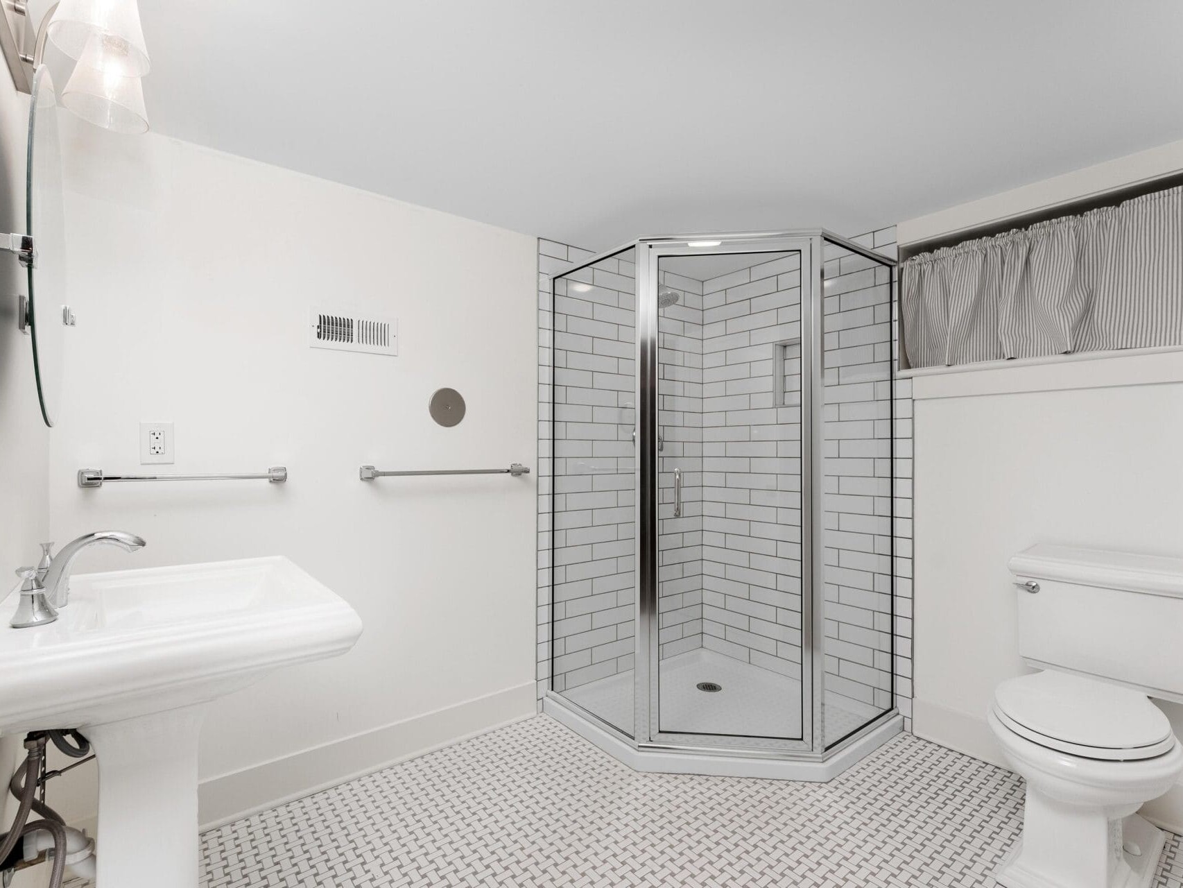 A modern bathroom with a white theme features a corner shower with glass doors and subway tile walls, a pedestal sink, and a toilet. The floor is tiled with a geometric pattern. A small, curtained window is above the toilet.