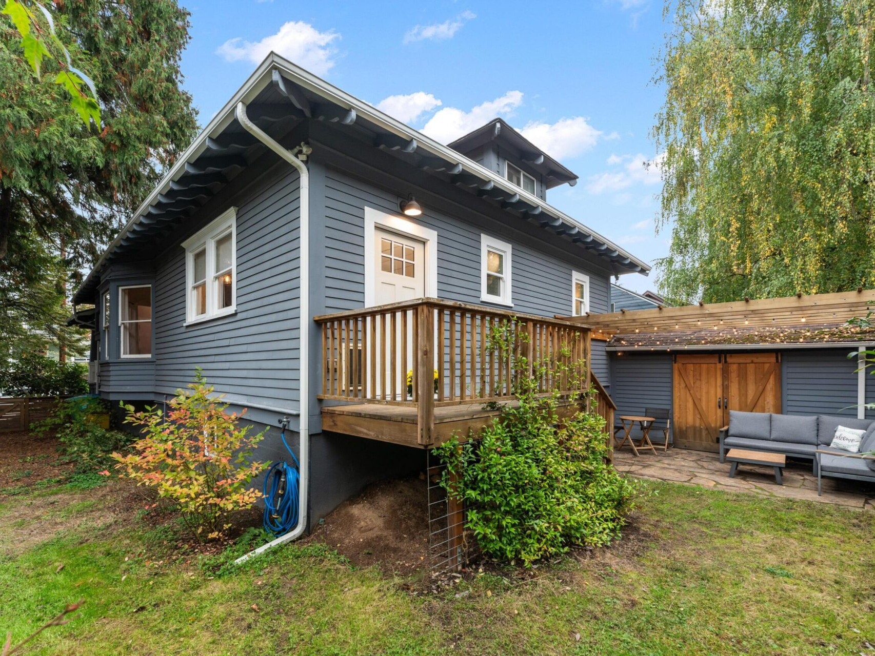 A charming blue-gray house with a small wooden deck and stairs leading to a lush green yard. The deck is surrounded by plants, and a cozy patio area with outdoor furniture is on the right. The sky is clear and blue.