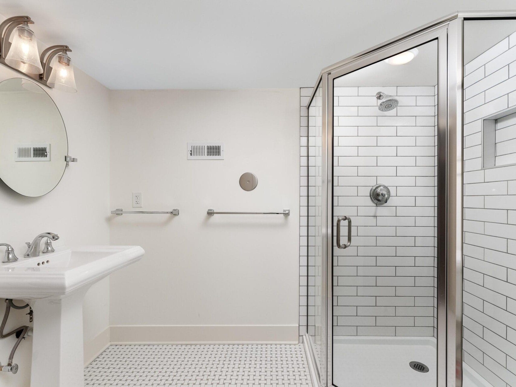 A modern bathroom with a white pedestal sink and round mirror on the left. A glass-enclosed shower with white subway tiles is on the right. The floor features patterned tiles, and two light fixtures illuminate the space.