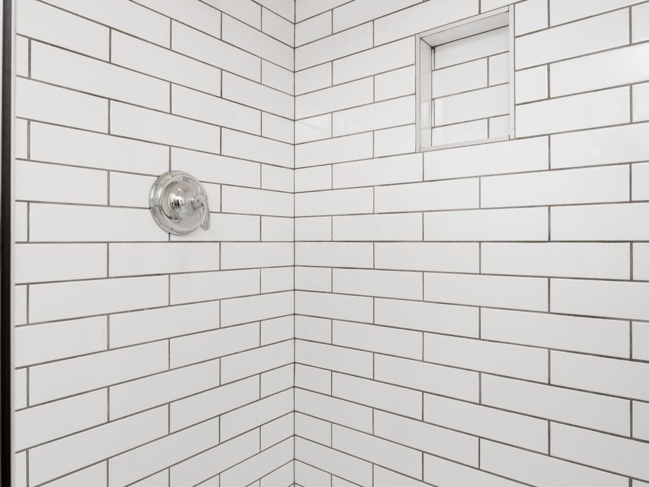 A shower with white subway tiles and dark grout, featuring a built-in wall niche and a silver showerhead. The floor is white with a central drain.