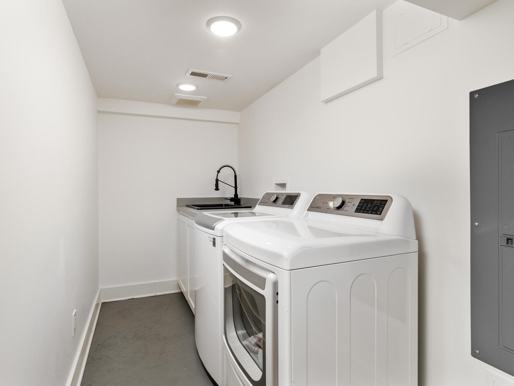 A clean, narrow laundry room with white walls features a washer and dryer side by side. Theres a black countertop with a sink and a black faucet to the left, under bright overhead lighting. Gray floors complete the minimalist look.