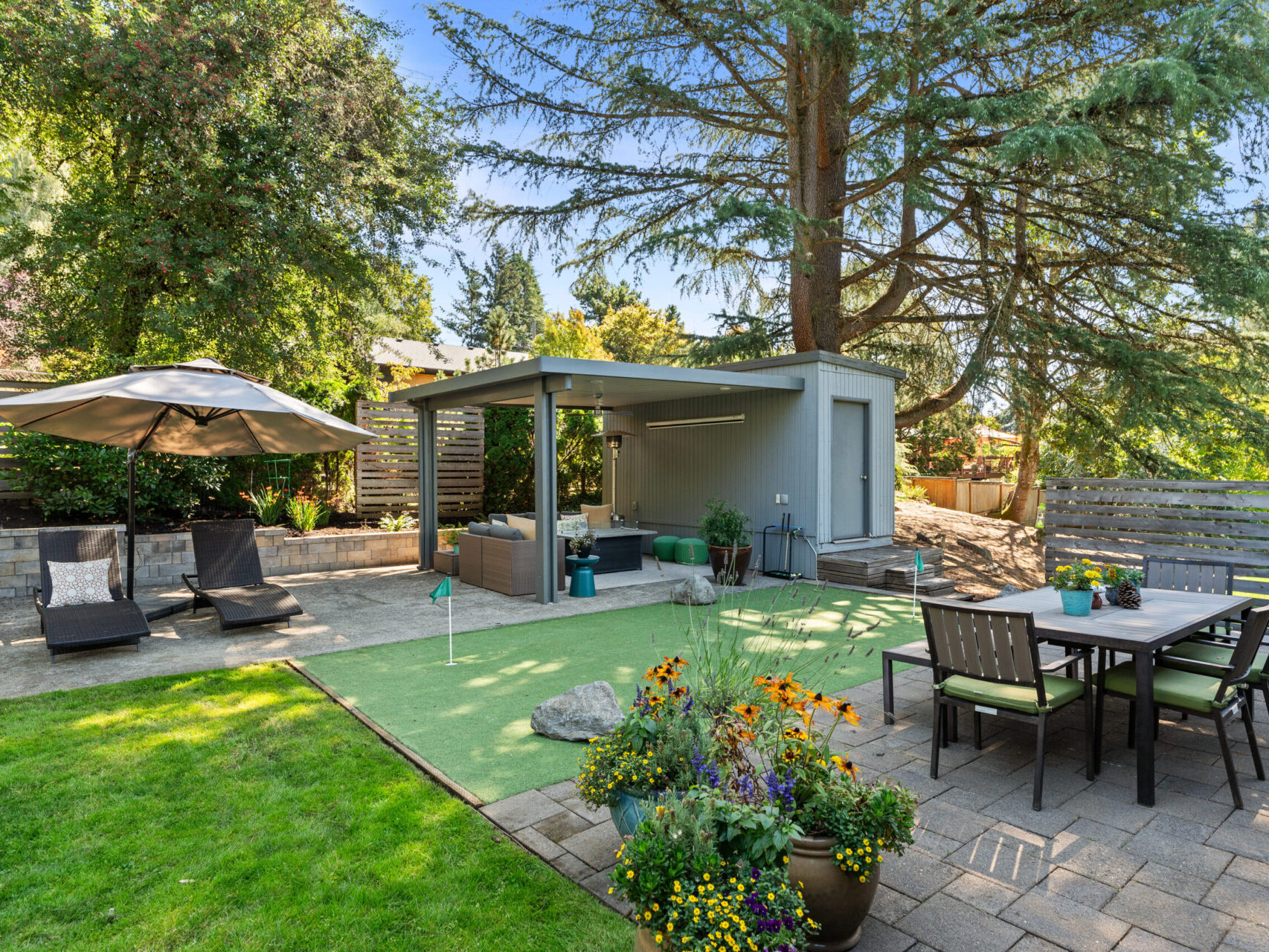 A backyard with a patio featuring a dining table and chairs, a small golf putting green, an outdoor seating area under an umbrella, a modern shed, and lush greenery including a large tree and various plants.