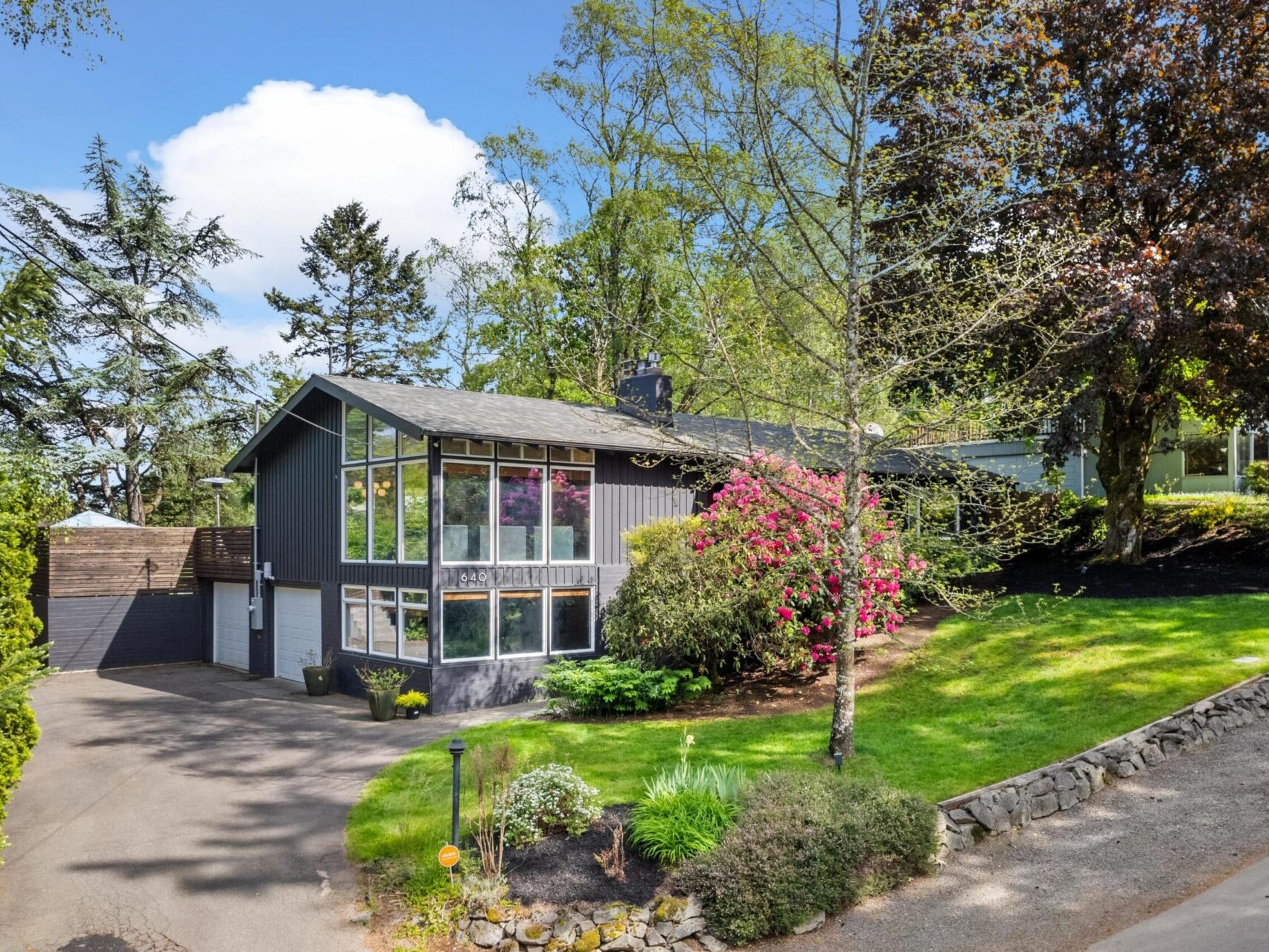 A modern two-story house with large windows and a dark exterior surrounded by lush greenery and blooming shrubs. The steep driveway leads to a garage, and the lawn is bordered by a stone retaining wall. Tall trees stand in the background under a blue sky.