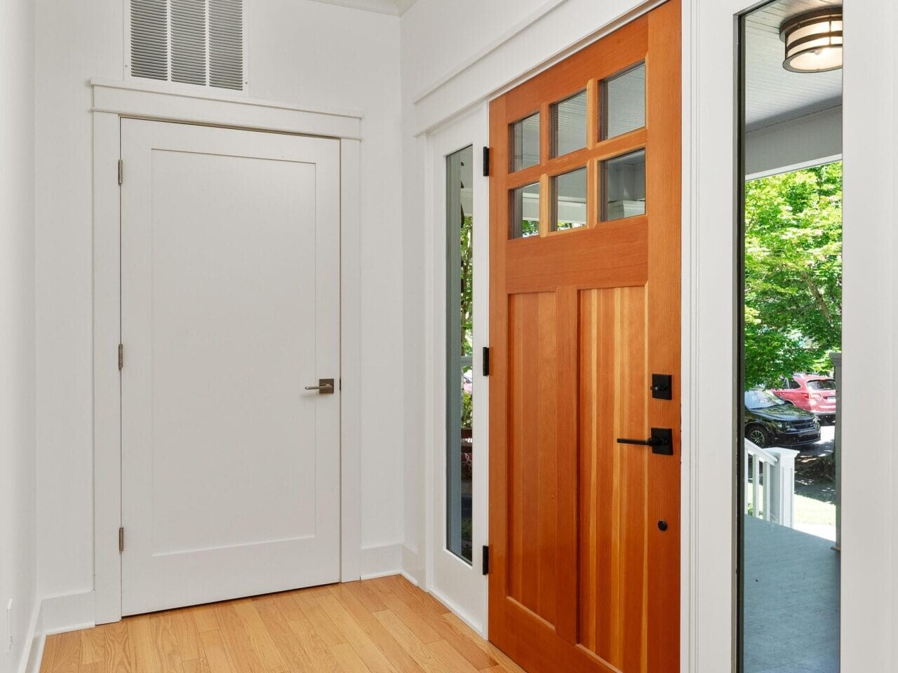 A bright hallway with light wood flooring features a wooden front door with glass panels, a white interior door, and a modern ceiling light. Large windows on the door reveal green trees outside.