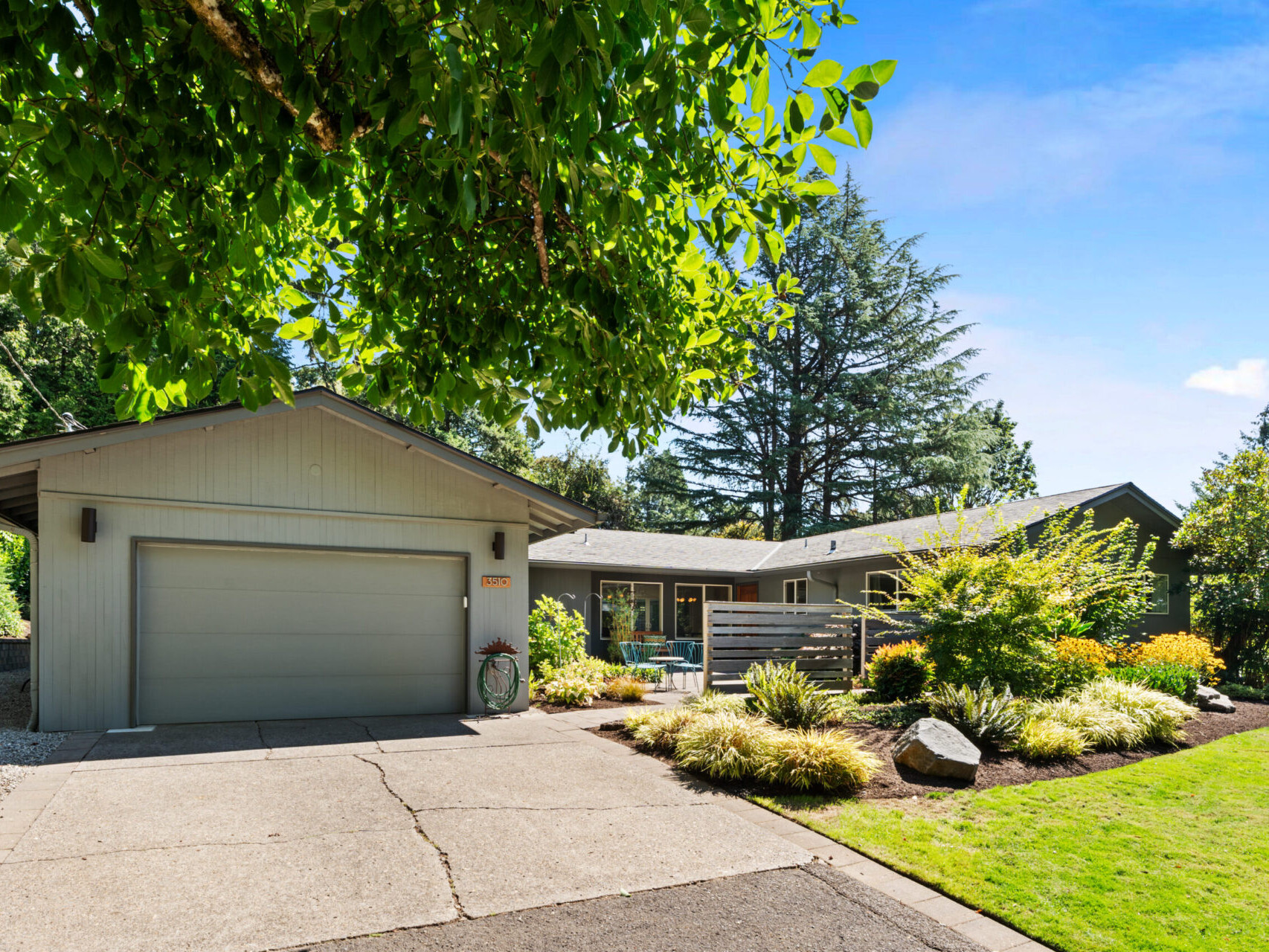 A single-story house with a large driveway and garage, surrounded by lush greenery and well-maintained landscaping. Theres a garden with various plants and a large tree in the front yard under a bright blue sky.