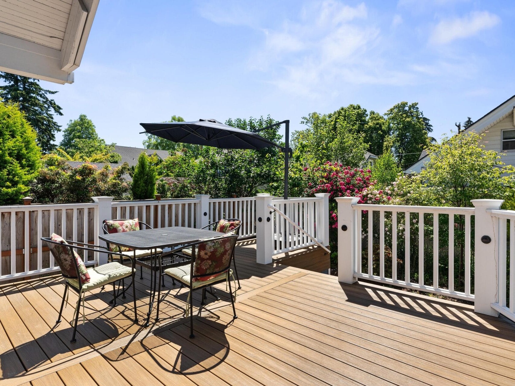 A sunny outdoor deck with wooden flooring, surrounded by a white railing. A black metal table and chairs are set up with floral cushions, partially shaded by a black umbrella. Lush greenery with blooming flowers and trees is in the background.