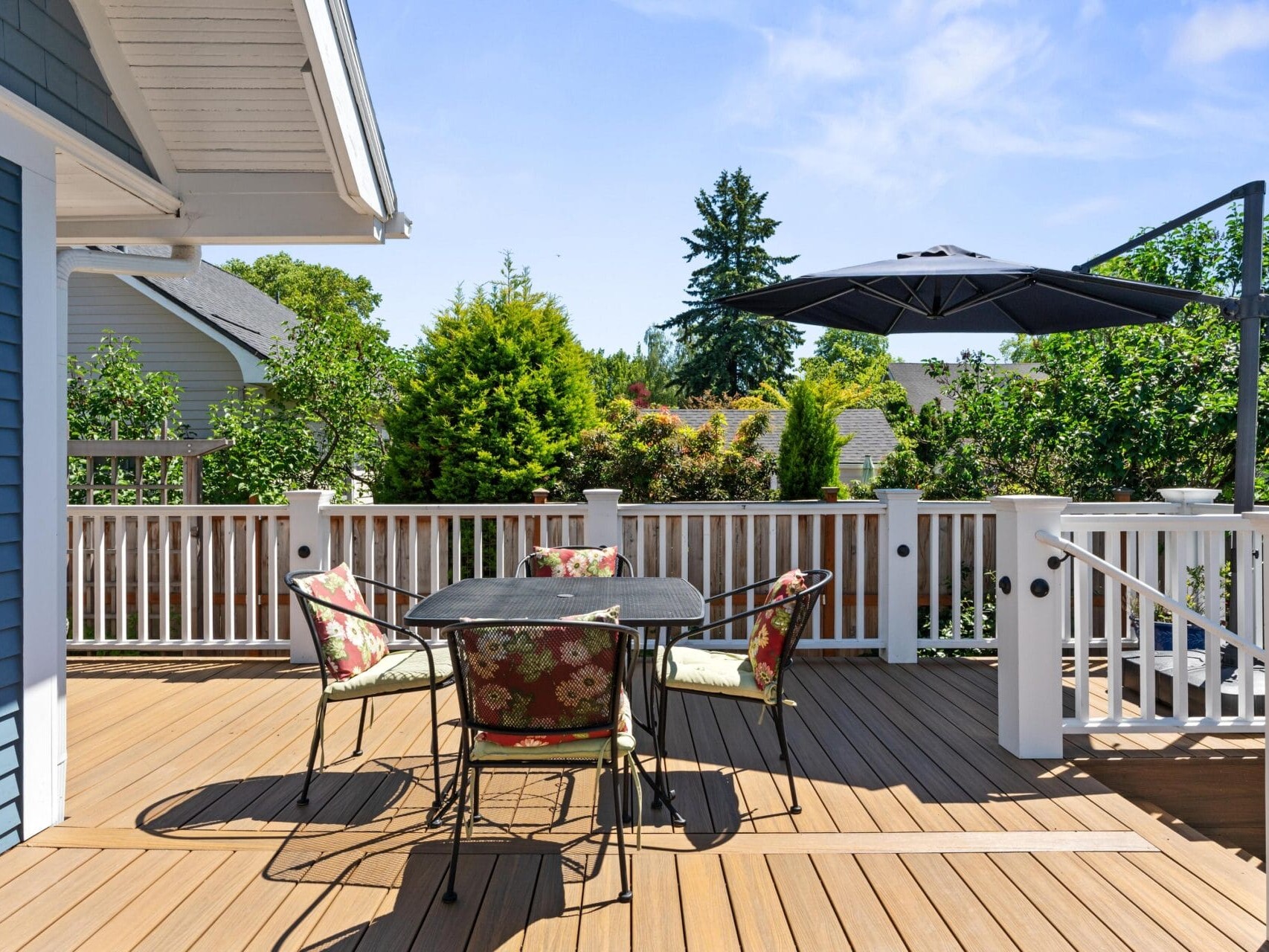 Outdoor wooden deck with a black metal table and four floral cushioned chairs. A large black umbrella provides shade. White railings surround the deck, and lush greenery is visible in the background under a clear blue sky.