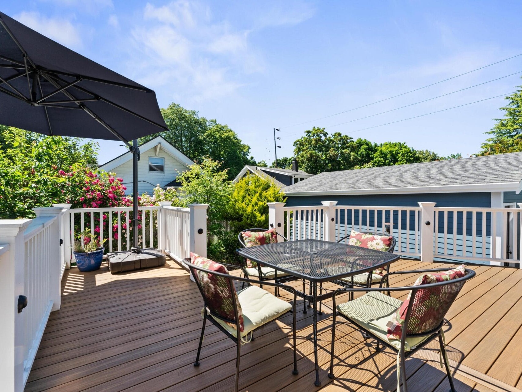 A sunny outdoor patio with a black umbrella, a metal table, and four chairs with floral cushions on a wooden deck. The patio is surrounded by white railings, lush greenery, and houses in the background.