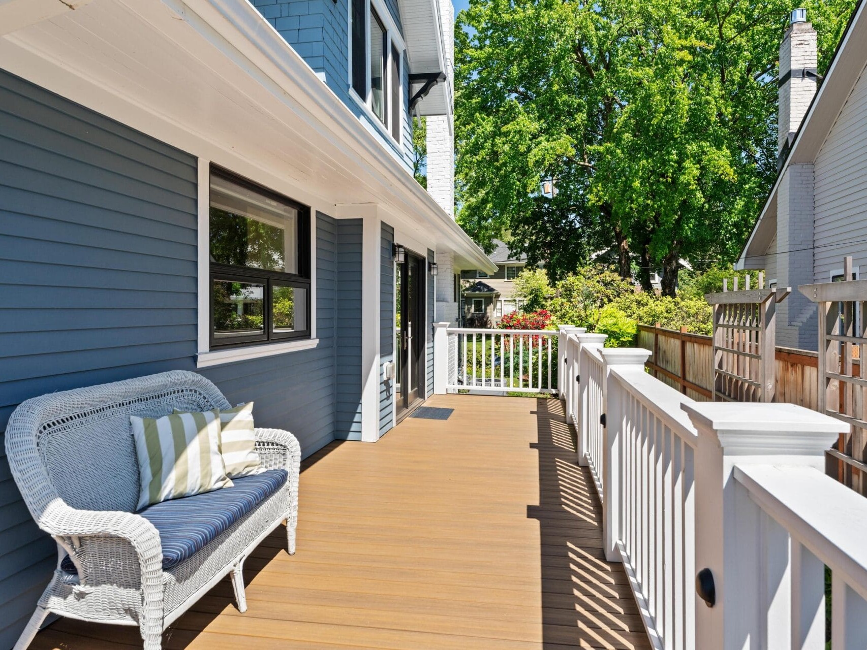 A sunlit wooden deck with a white railing. A wicker loveseat with blue and white striped cushions sits near a blue house wall. Lush greenery and trees are visible beyond the railing, creating a serene outdoor space.