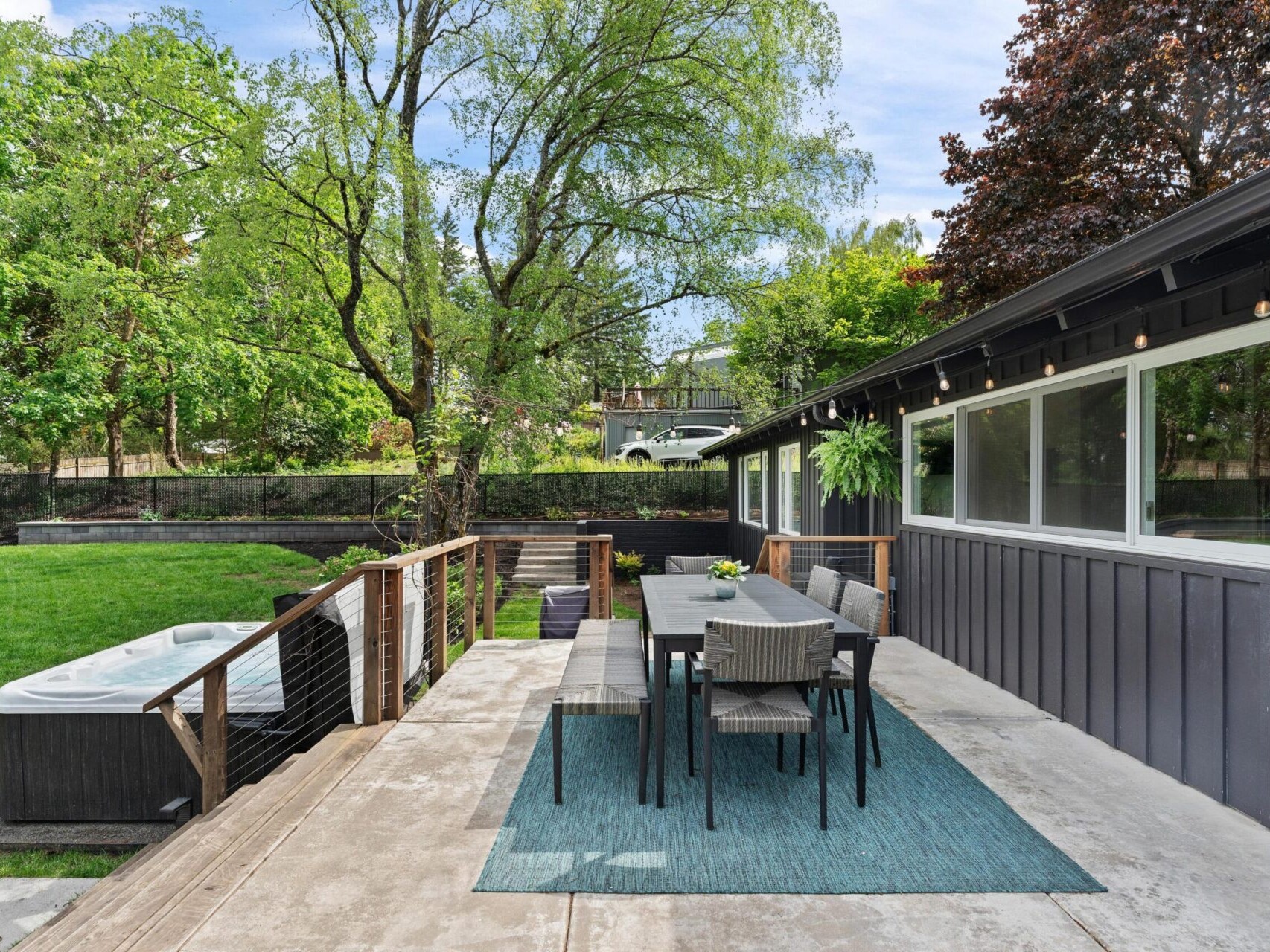 A modern outdoor patio featuring a dining table with chairs on a blue rug, overlooking a lush green lawn. A hot tub is situated nearby, surrounded by trees and greenery. The house exterior is dark with large windows.