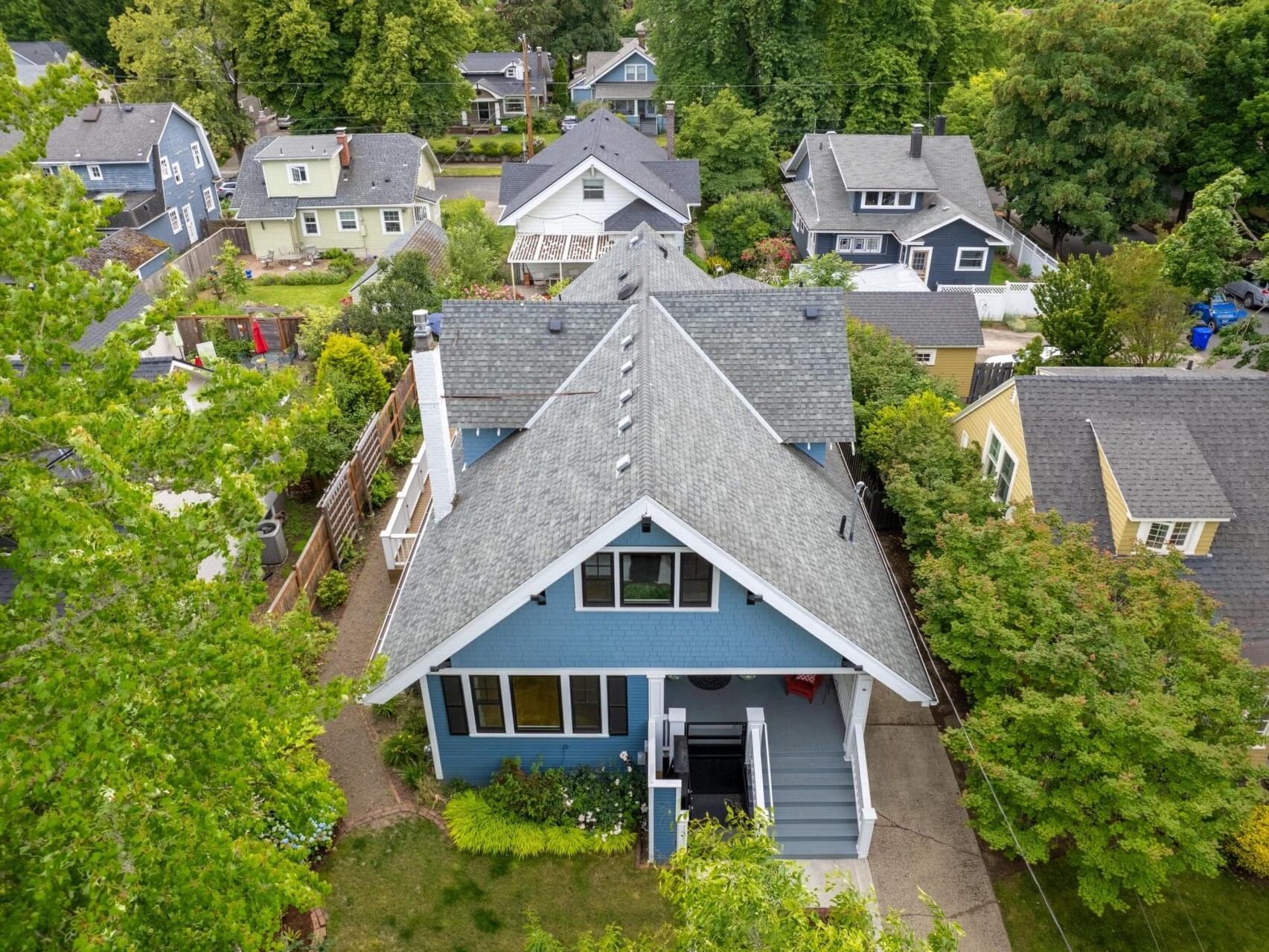 Aerial view of a residential neighborhood with a central blue house surrounded by trees. The house features a gray roof and front steps leading to the entrance. Other houses with similar architecture are visible in the background.