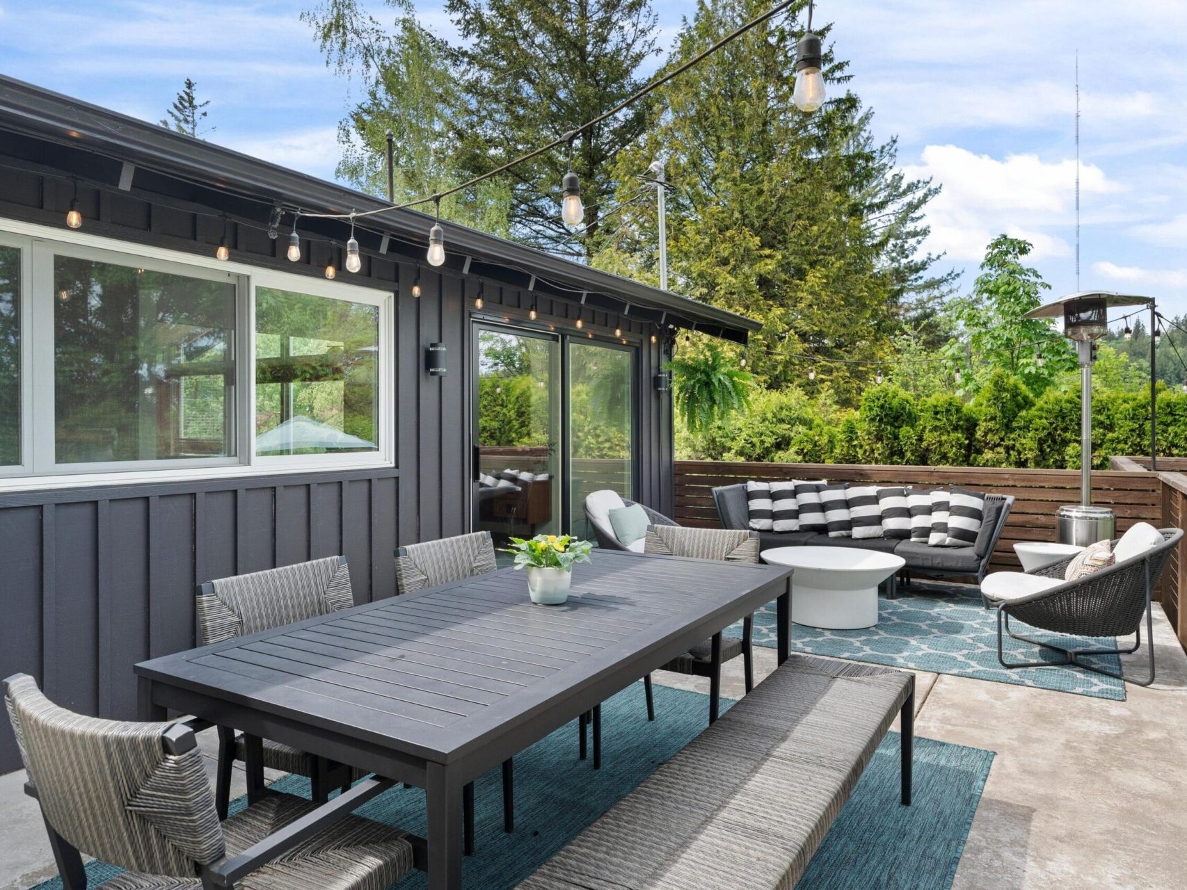A stylish outdoor patio features a long dining table with chairs and a bench. String lights hang above, and a cozy seating area with a sofa and chairs is arranged on a blue rug. Tall trees and clear skies provide a serene backdrop.