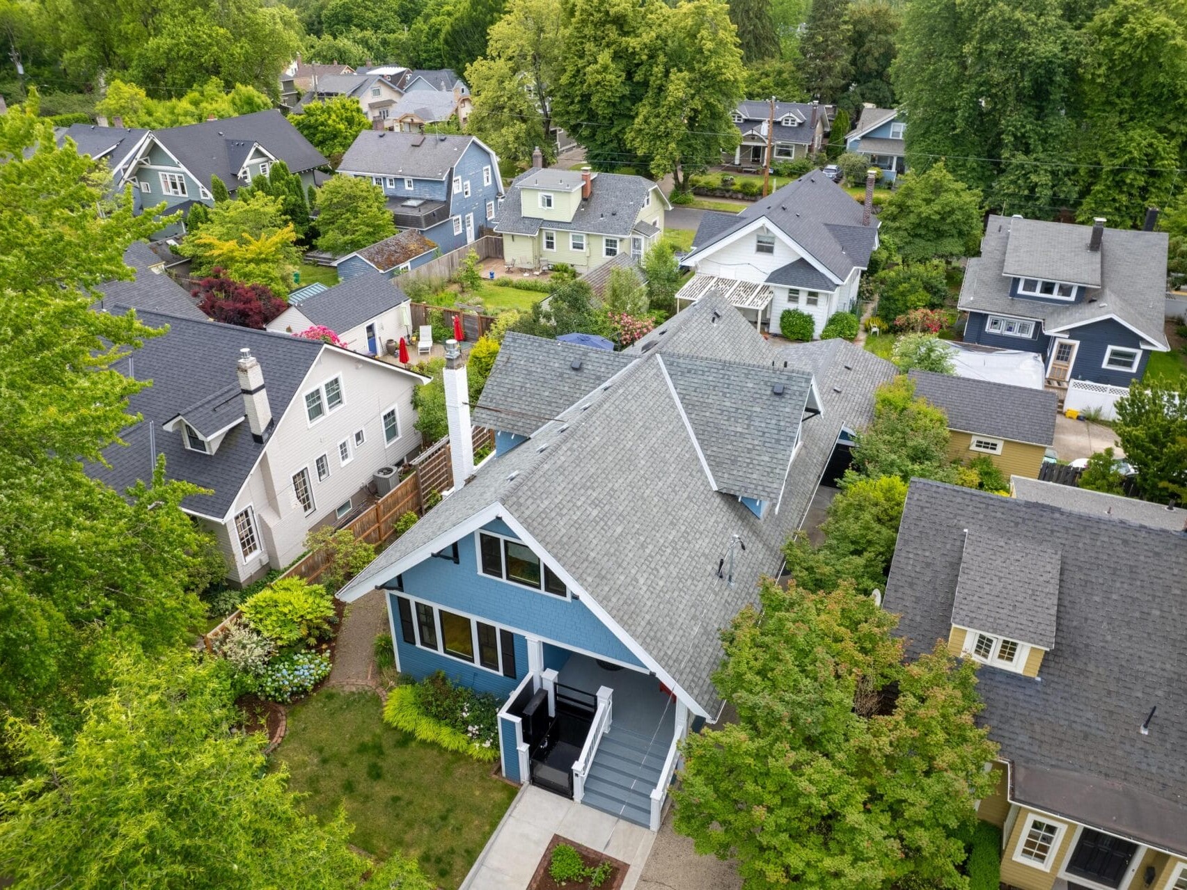 Aerial view of a residential neighborhood with several houses surrounded by lush green trees. The homes have sloped roofs and are painted in various colors like blue, white, and gray, creating a picturesque and serene suburban setting.