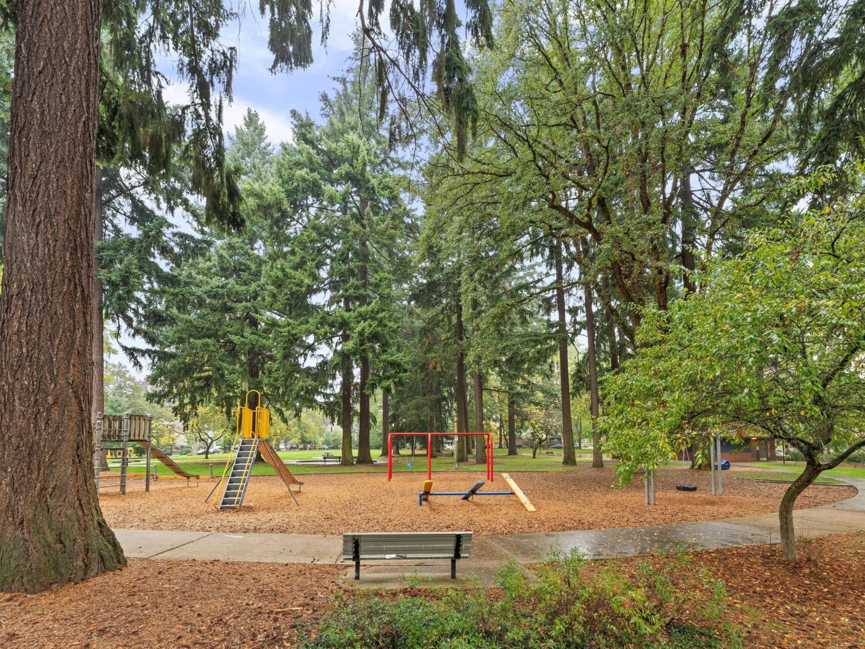 A park with tall trees features a playground area. There are swings, a yellow slide with a blue ladder, and a seesaw. The ground is covered with wood chips, and a green bench is placed nearby. Paths wind through the area surrounded by lush greenery.