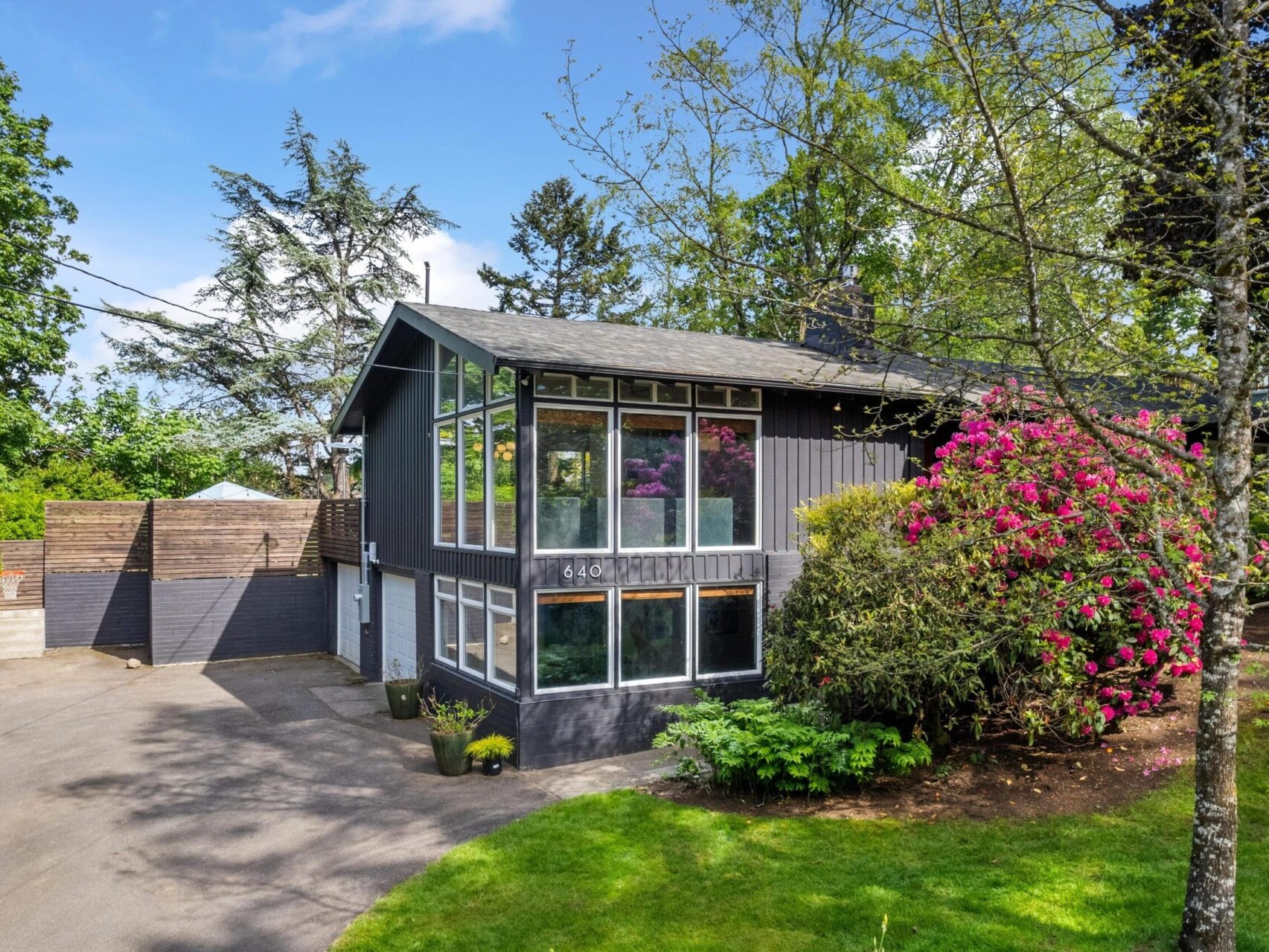 A modern two-story house with large windows, surrounded by lush greenery and vibrant pink flowers. A driveway leads to a garage, and trees frame the property under a clear blue sky.