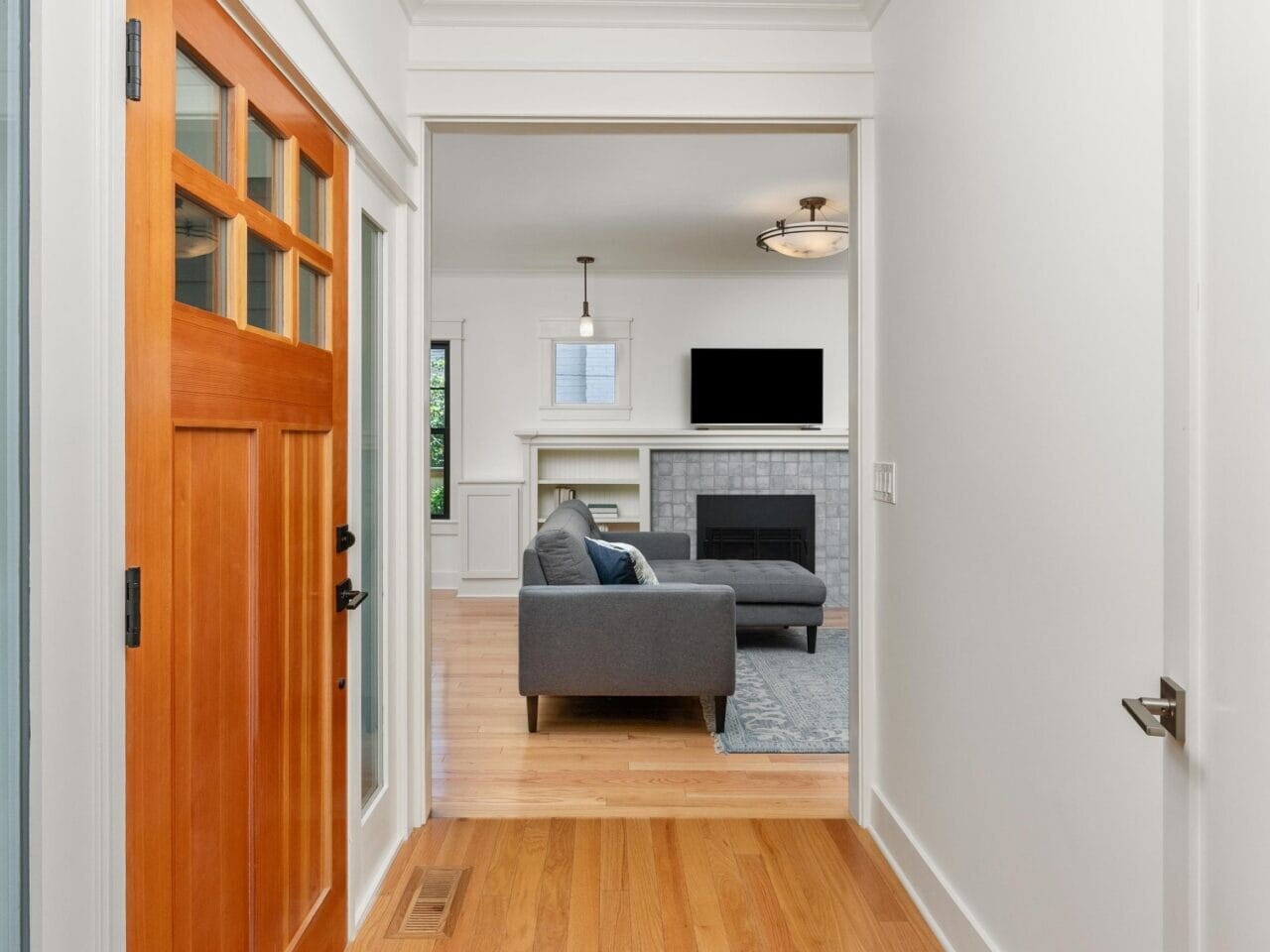 View of a modern homes hallway with a wooden front door on the left. Light wood flooring leads to a living room featuring a gray sofa, a fireplace, and a wall-mounted TV. The rooms are bright with white walls and ceiling lights.