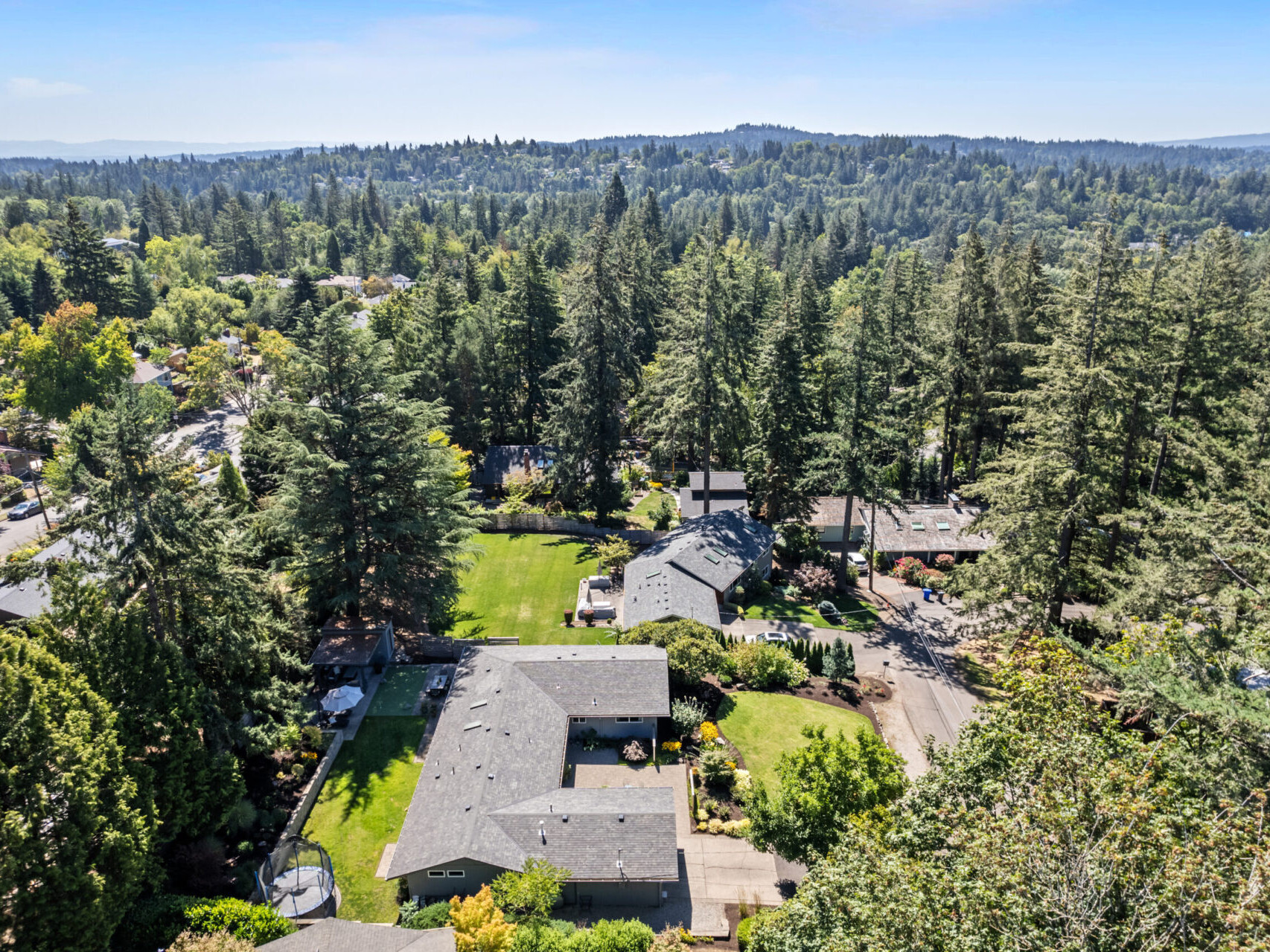 Aerial view of a suburban neighborhood surrounded by dense green trees and hills. Houses have spacious yards with bright green lawns. A clear blue sky is visible in the background.