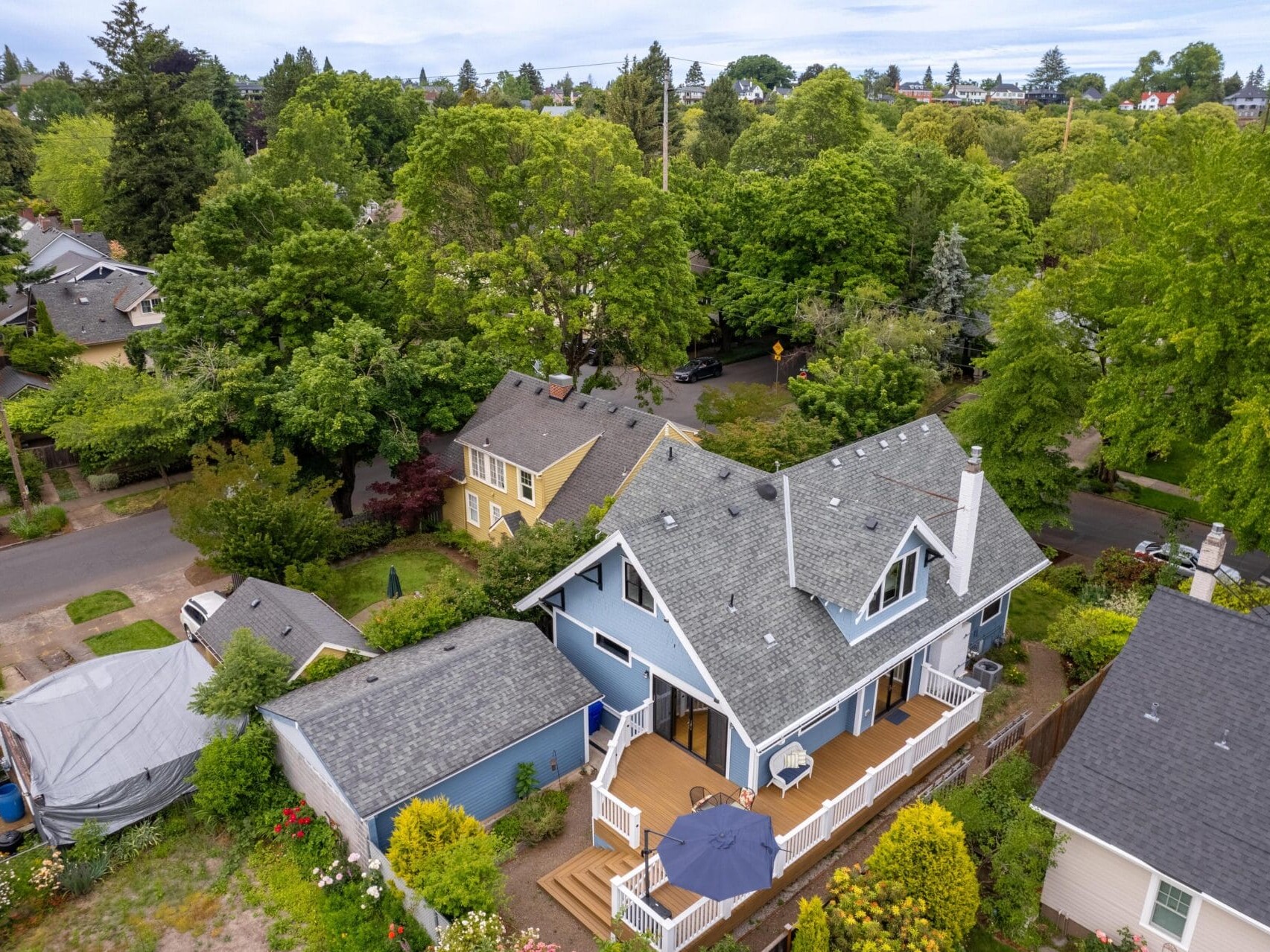 Aerial view of a residential neighborhood with lush greenery and trees. Central focus on a blue house with a gray roof and wooden deck. Adjacent houses are visible, surrounded by vibrant gardens and landscaped yards.