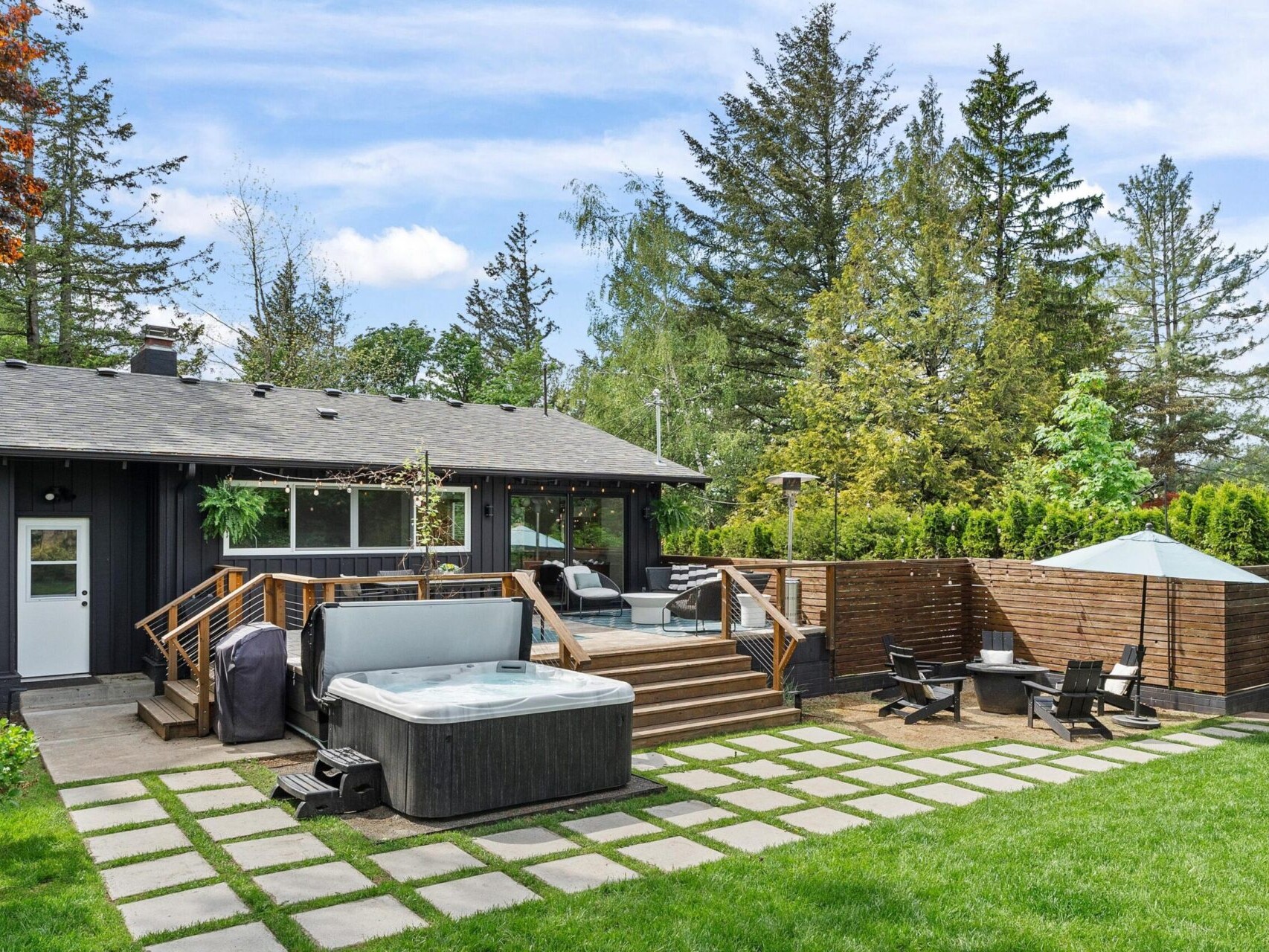 A modern backyard with a black house, wooden deck, and hot tub. There are wooden privacy screens, lounge chairs, and a fire pit area. The yard features a grassy lawn, stepping stones, and is surrounded by tall trees and greenery.