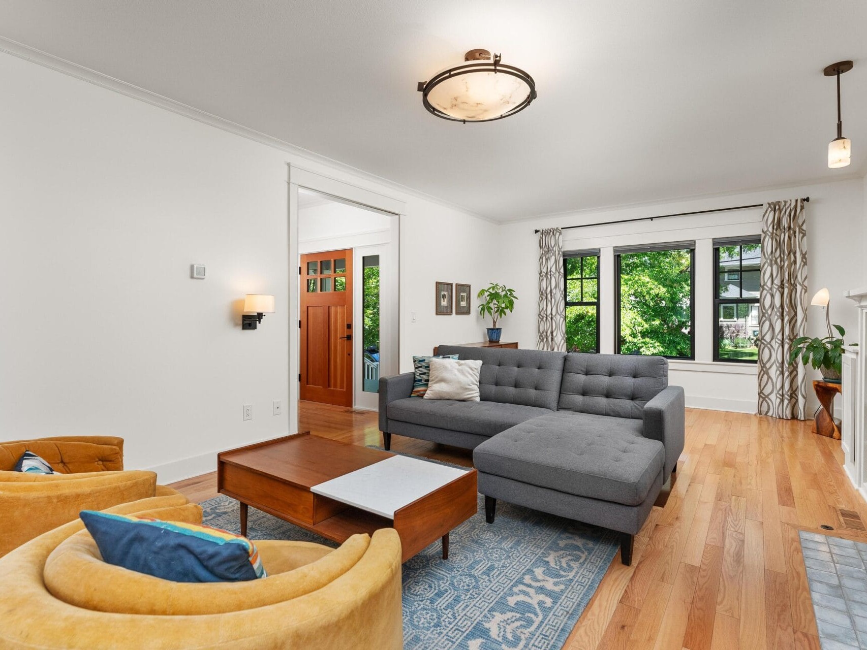 A cozy living room features a gray sectional sofa, two mustard-yellow armchairs, and a wooden coffee table on a blue patterned rug. Large windows with curtains let in natural light, highlighting the wooden floor and minimalistic decor.
