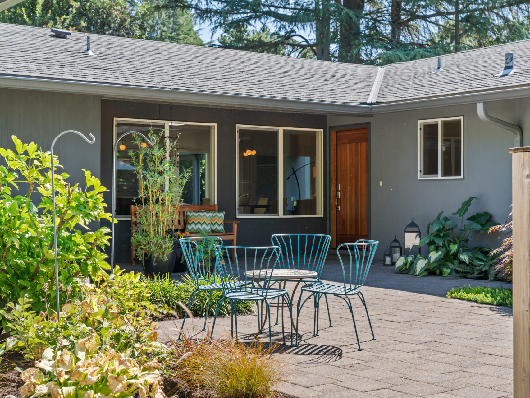 A cozy courtyard with a turquoise metal patio set on a paved area. Surrounding greenery and plants enhance the serene atmosphere. Large windows and a wooden door are visible on the gray exterior of the building.
