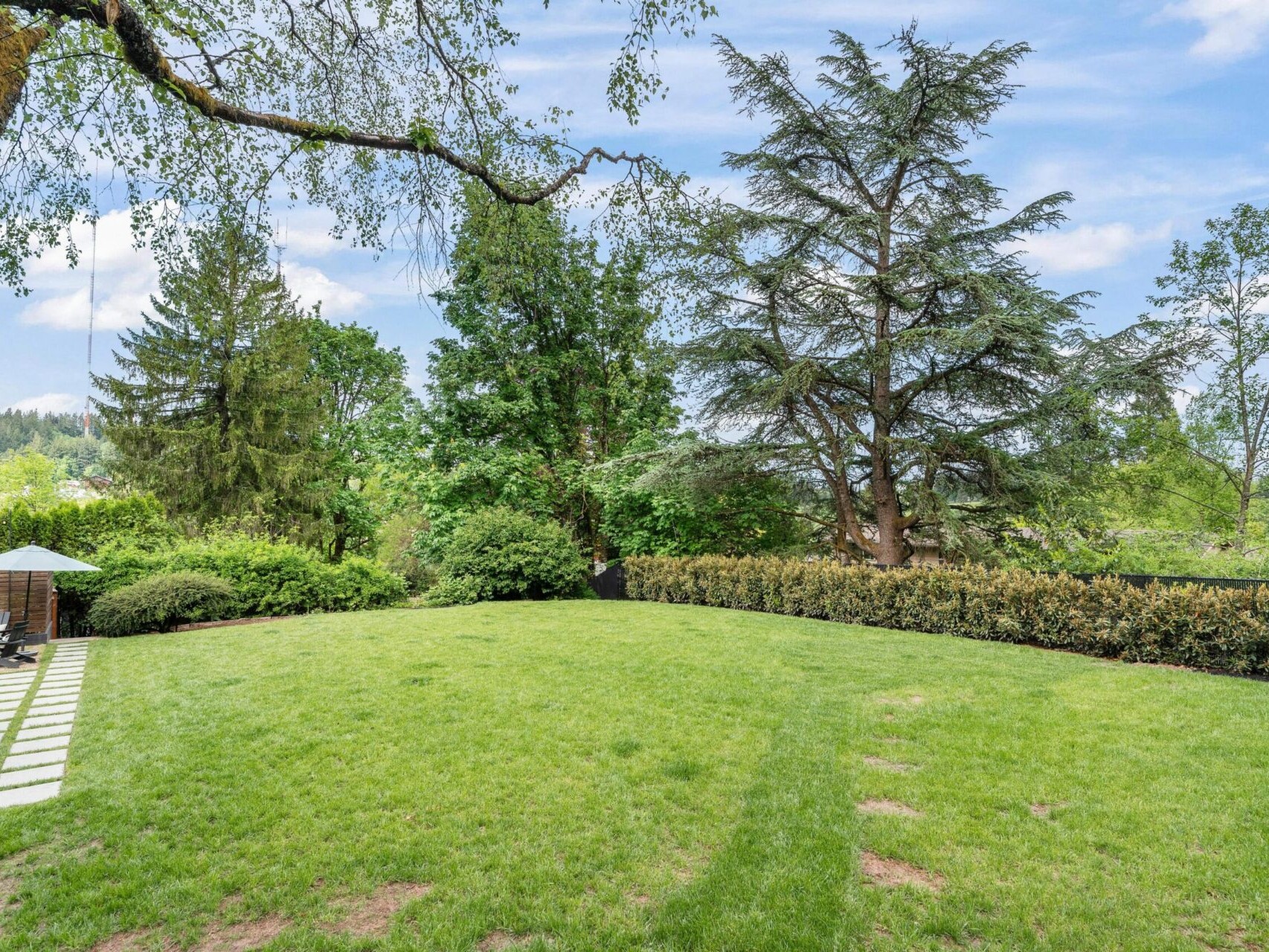 A spacious, lush green backyard featuring a well-maintained lawn, tall trees, and a small patio area with outdoor furniture under an umbrella. A stone path leads from the patio, surrounded by dense shrubs and trees against a clear blue sky.