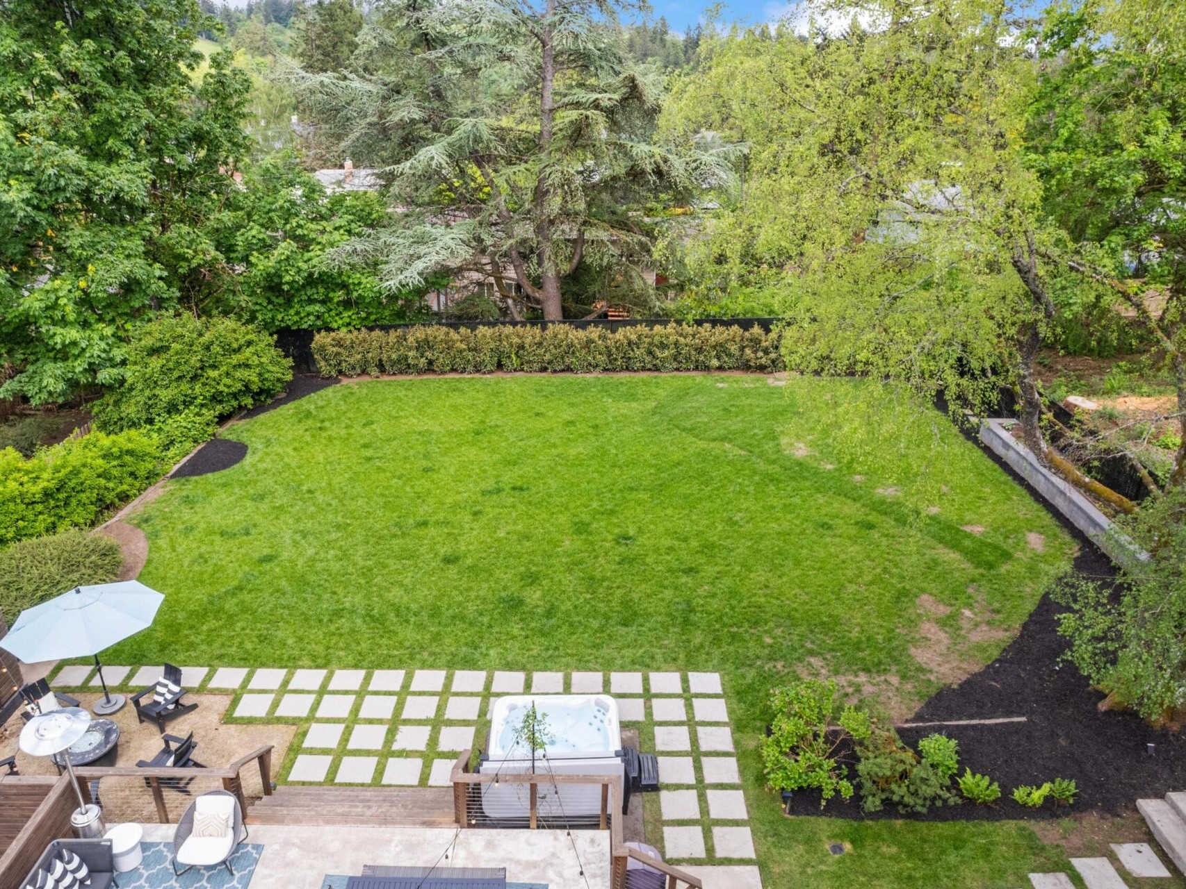 Aerial view of a spacious backyard featuring a lush green lawn, surrounded by trees and bushes. The foreground includes a patio area with a hot tub, lounge chairs, and a table with an umbrella, with stepping stones leading to the yard.
