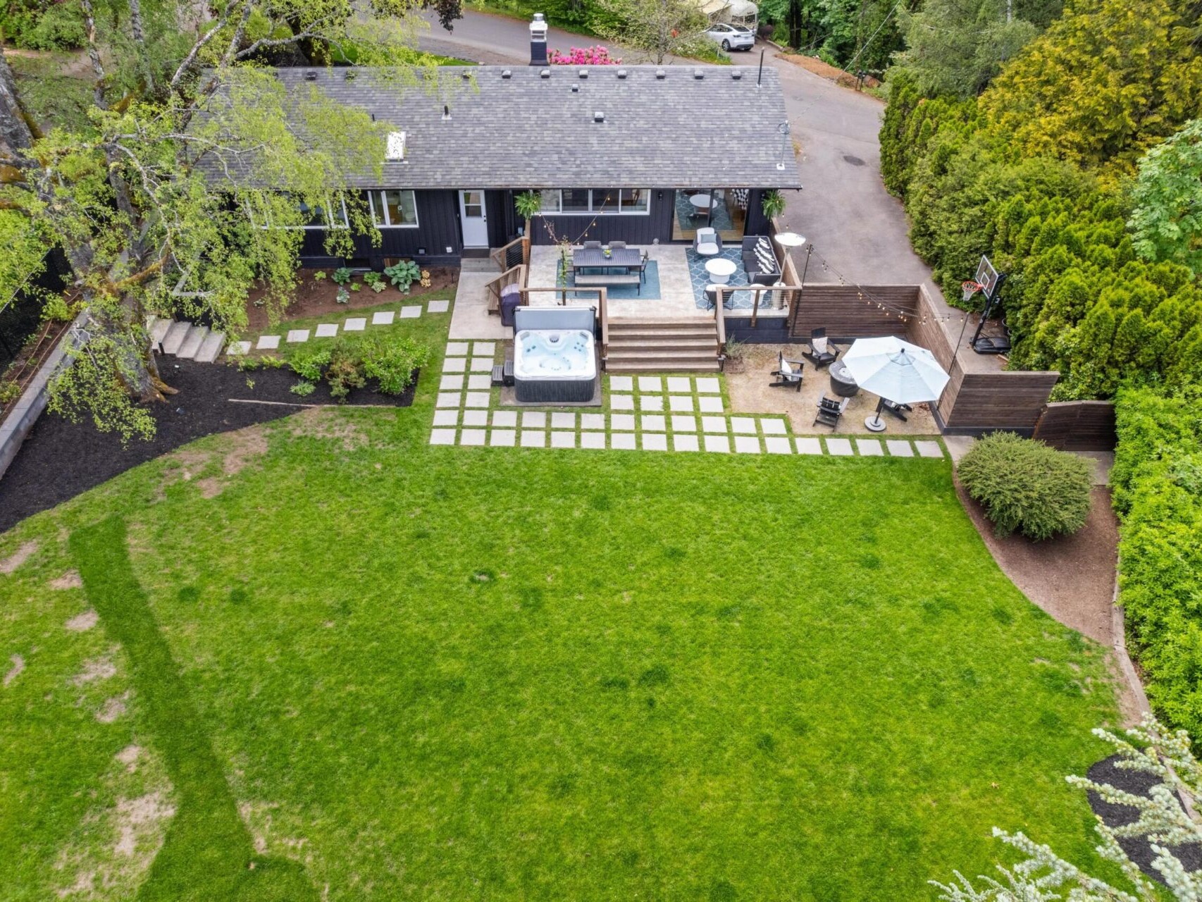 Aerial view of a house with a large backyard. The yard features a patio with a hot tub, outdoor furniture, and a pathway. Surrounding the yard are trees and shrubs, with a paved driveway leading to the house.