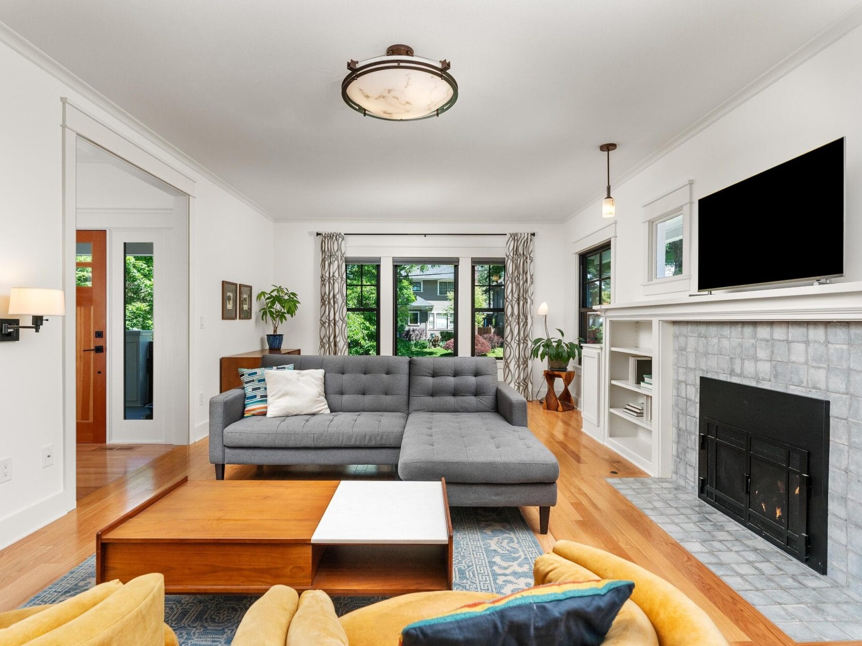 A modern living room with a gray sectional sofa, colorful pillows, and a wooden coffee table. A fireplace with a mounted TV is on the right, flanked by shelves. Large windows offer a view of greenery outside, and the room features wood floors.