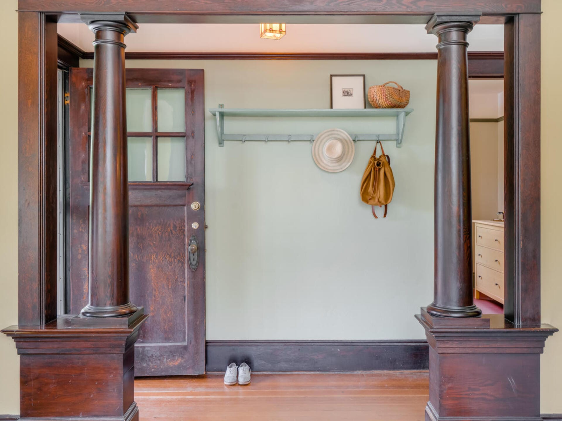 Entryway with wooden columns framing a door, a light green wall with a shelf and hooks, holding a bag and hat. Below, a pair of shoes is placed neatly. A wooden door with glass panes completes the cozy interior.