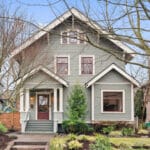 A charming two-story house with a gray exterior and white trim, surrounded by bare trees and lush landscaping. The home features a covered front porch and multiple windows, creating a welcoming appearance. A clear sky is visible in the background. 