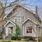 A large, green, two-story house with a gabled roof and a covered porch. The home is surrounded by bare trees and trimmed shrubs, and features multiple windows and a brick chimney against a clear blue sky. 