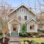 A charming two-story green house with white trim, surrounded by leafless trees and landscaped shrubs. The front porch includes a small staircase leading to the entrance. The sky is clear, indicating its a bright, sunny day. 