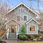 A charming two-story house with green wooden siding and a gabled roof, surrounded by leafless trees and various shrubs. The house features a front porch with steps leading to a maroon door and is set on a neatly landscaped yard. 