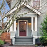 Front view of a two-story house with pale green siding and white trim. There are steps leading to a maroon front door, framed by columns. Trees and shrubs decorate the front yard, and a wooden fence is visible on the left. 