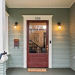A front porch with green siding features a red door with glass panels. A Hooray! doormat lies at the entrance. Two wall-mounted lanterns illuminate the space, and the house number 2426 is displayed above the door. 