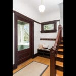 A well-lit foyer with wooden stairs, white walls, and dark wood trim. There’s a small bench with throw pillows, and hooks above it. A window and glass-paneled door reveal a porch and greenery outside. A hanging light fixture illuminates the area. 