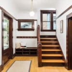 A hallway with wooden floors and trim features a staircase leading up. Theres a wall-mounted coat rack and shelf above a bench with pillows. A window is on the stairway wall. A pendant light hangs from the ceiling, illuminating the space. 