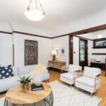 A cozy living room with hardwood floors, featuring a light gray sofa, two white chairs, and a round wooden coffee table. Art on the wall and a staircase in the background add to the elegant, inviting atmosphere. 