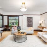 A bright, spacious living room with light-colored walls and hardwood floors. It features a white sofa, a round coffee table, a floor lamp, a large window with natural light, and a modern wall art piece. A cozy chair and bench complete the setting. 